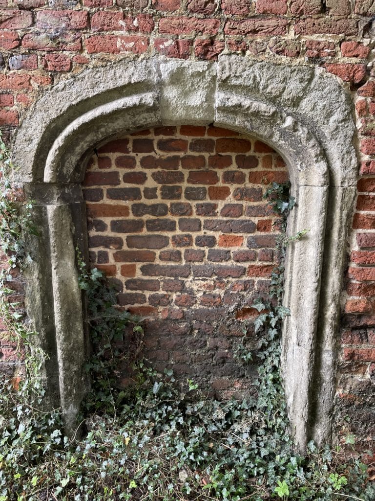 A bricked up, stone Tudor archway, partially buried in the earth and overgrown with ivy.