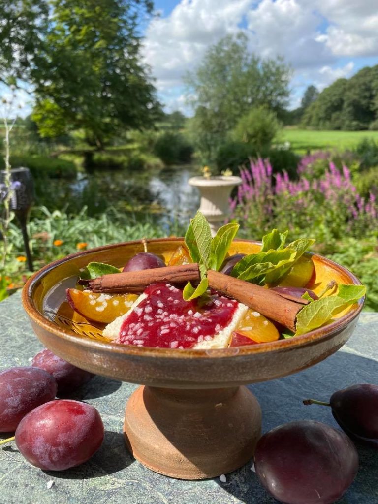 A picture of Tudor Plum Tart  on a pedestal platter in a garden with fresh plums around the base